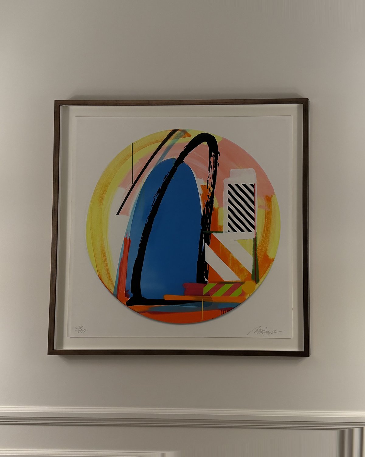 Large colourful circular abstract print in a slim bronze box frame with a wide white border, hung on a white panelled wall.