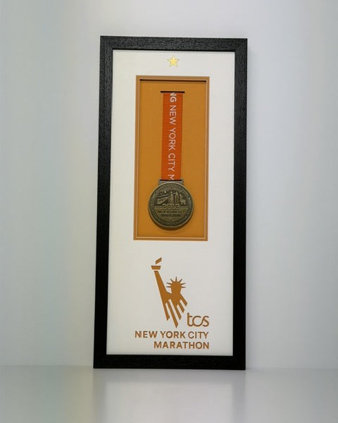 Black frame holding a New York City Marathon finisher's medal on an orange ribbon, with the marathon logo cut into a white mount.