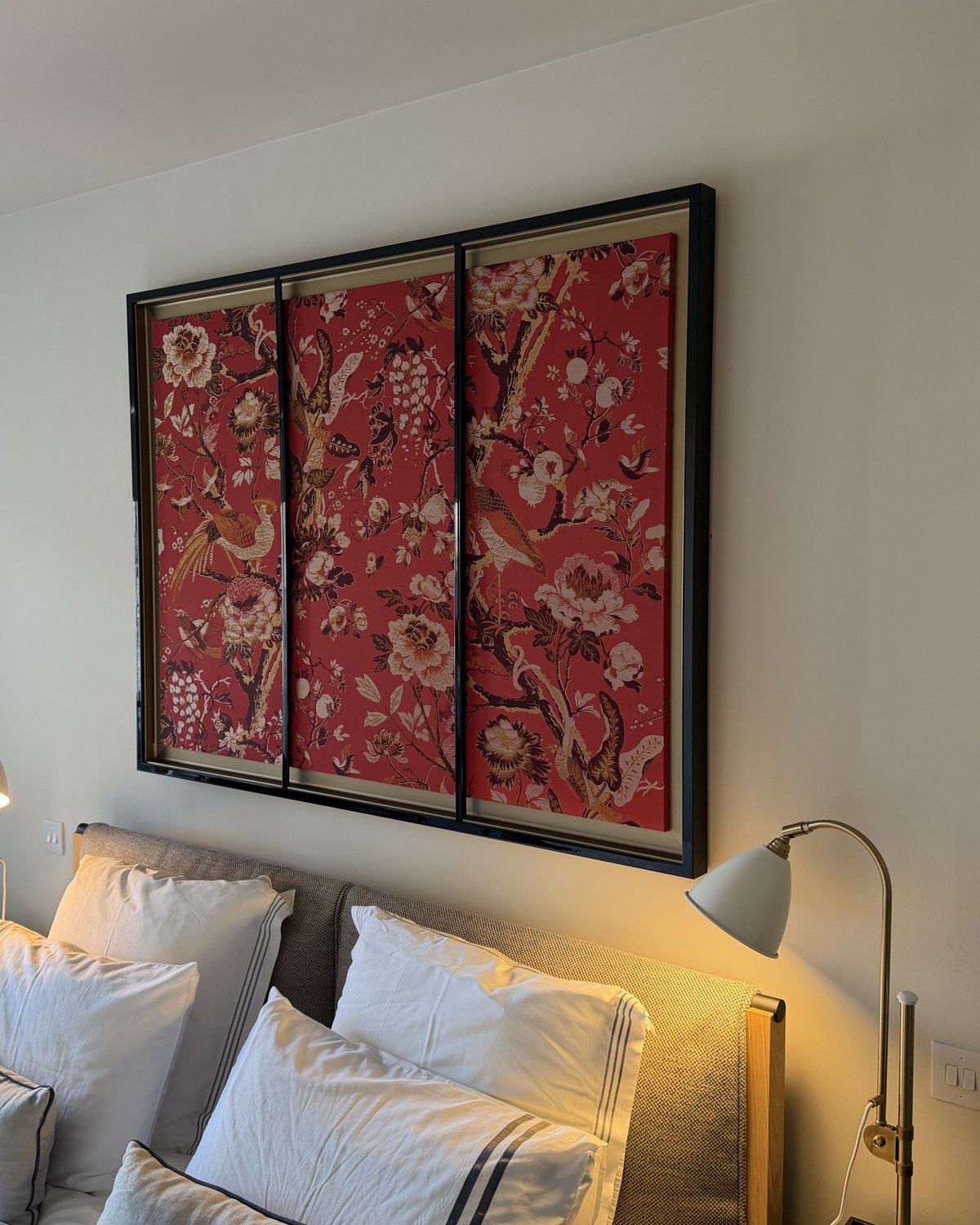 Three red floral fabric panels in a black frame with gold backing, hung above a bed.
