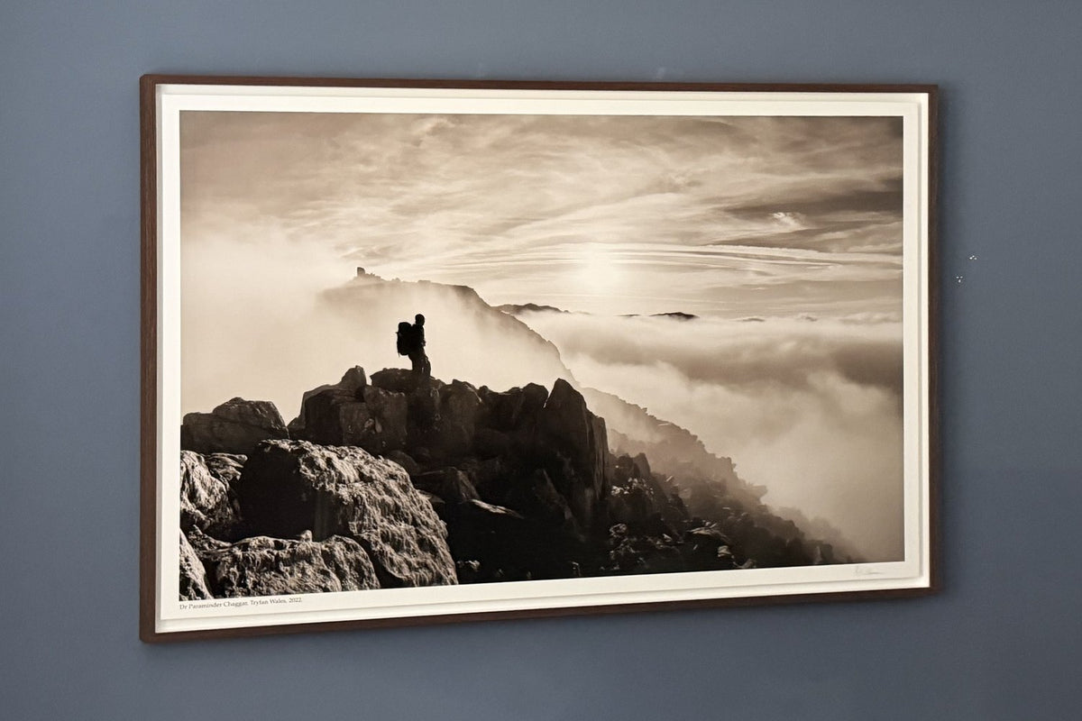Large sepia photograph of a walker silhouetted on a rocky Welsh summit above the clouds, in a dark timber frame with a white border, on a blue wall.